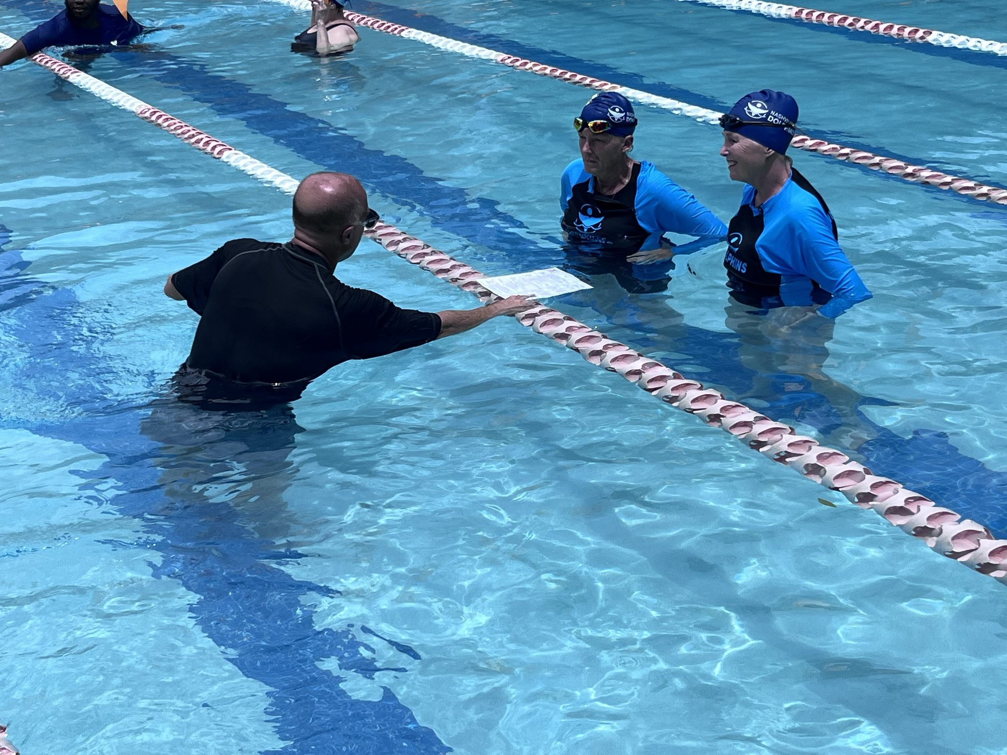 Bill Brenner in the water reviews a handout with two adult swimmers at the lane line